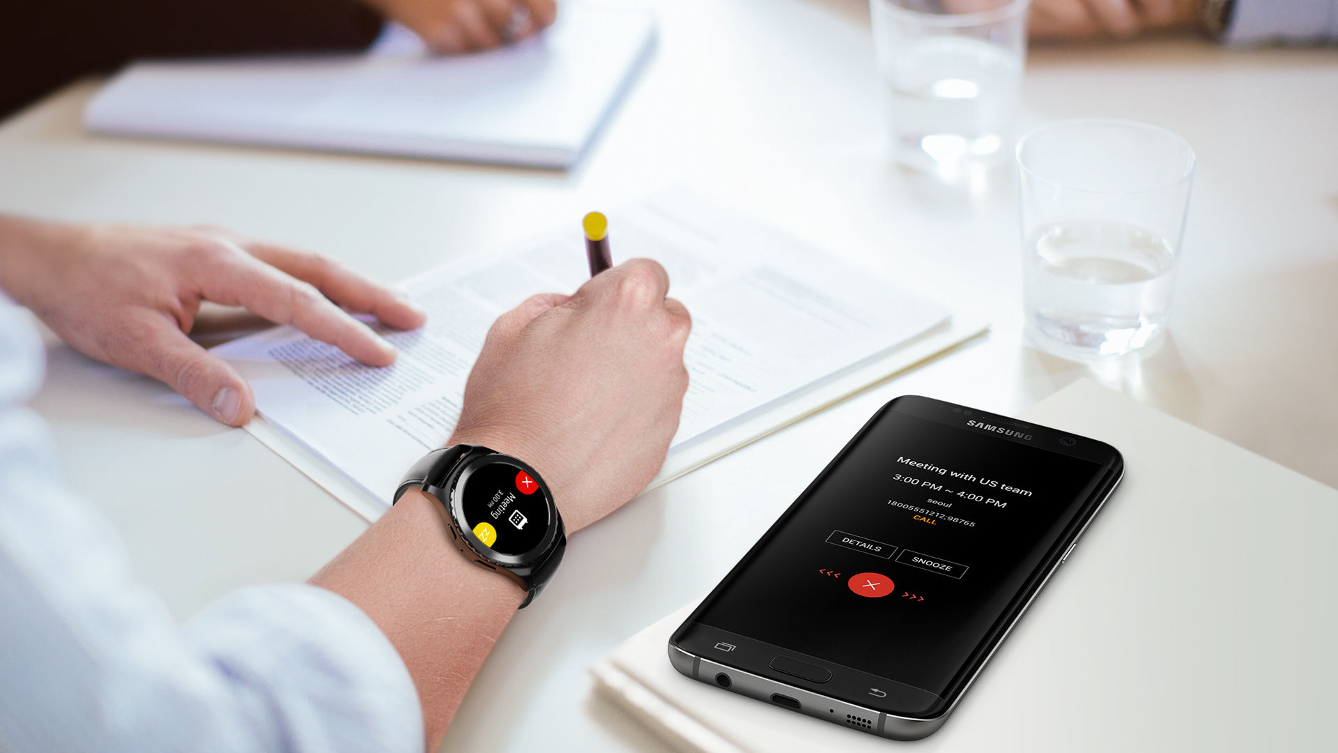 An image showing an office worker writing on a document. On his wrist is a smart watch that is receiving a conference call notification from a paired Galaxy S7 edge device, which is shown beside the worker, with a conference call notification on its screen.